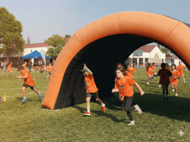 Elementary students in orange shirts running through an inflatable arch at an Apex fun run