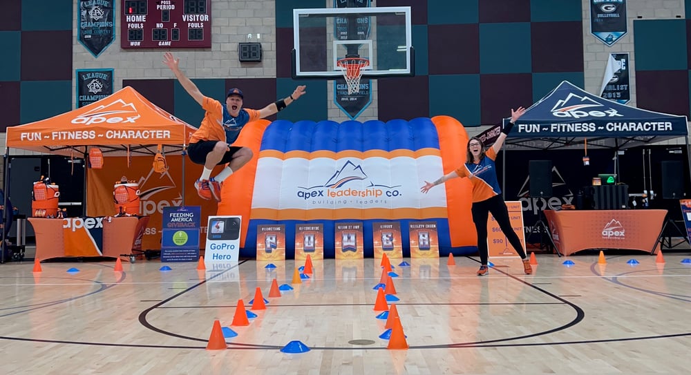 Apex coaches jumping in front of the Apex inflatable and obstacle course in a school gym